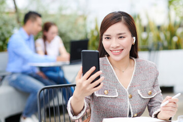Obraz premium Positive young Vietnamese businesswoman sitting at table in outdoor cafe and having video call with coworker