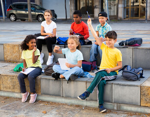 Fototapeta premium Focused diligent schoolchildren sitting with hands raised during lesson outside school in warm autumn day.