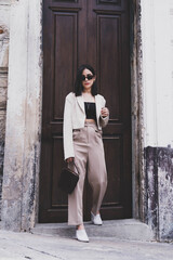 Asian pretty young hipster woman in trendy sunglasses in a vintage jacket in brown long pants with a bag stands near a white building in the city. Beautiful girl for a walk. Stylish women's clothing.