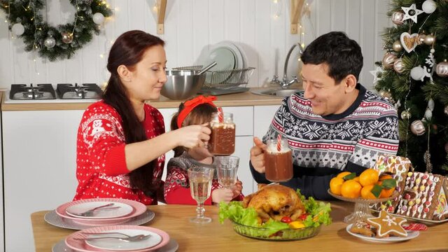 happy husband and wife drink tasty beverages sitting at festive dinner with little daughter by Christmas tree at home