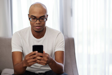 Obraz na płótnie Canvas Smiling young Black man in glasses sitting on sofa and checking news on social media
