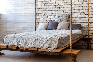 Wooden bed with pillows and a fur blanket hanging on a ropes in loft interior of a stylish modern bedroom.