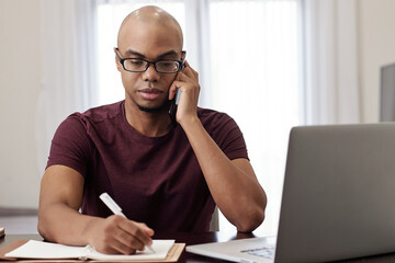 Portrait of serious young businessman calling on phone and taking notes in planner when talking on...