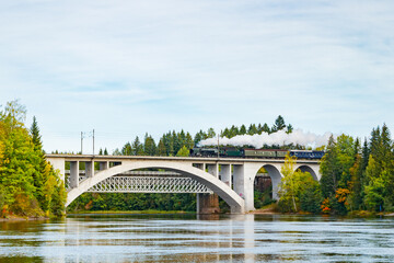 Naklejka premium Kouvola, Finland - 18 September 2020: Autumn landscape of bridge with moving old steam passenger train Ukko-Pekka and Kymijoki river waters in Finland, Kouvola, Koria