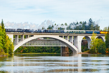 Fototapeta premium Kouvola, Finland - 18 September 2020: Autumn landscape of bridge with moving old steam passenger train Ukko-Pekka and Kymijoki river waters in Finland, Kouvola, Koria