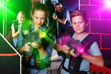 Happy teen brother and sister with laser pistols playing laser tag in dark labyrinth with their parents
