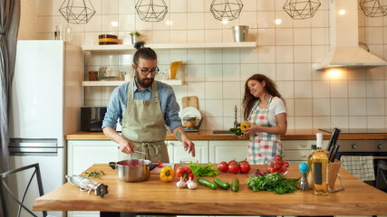 Italian cook cutting, preparing vegetables for cooking healthy meal. Young woman, girlfriend in apron looking at him, helping him in the kitchen. Hobby, lifestyle, relationships concept