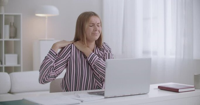 young woman is feeling sharp ache in neck and shoulder during work in home office, chatting online and writing in copybook