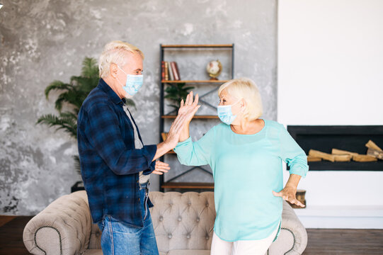 Joyful Senior Couple With A Medical Mask On Their Faces Has Fun Together At Home During Pandemic, Quarantine. A Mature Man And Senior Woman Wearing Protective Masks Dancing In The Living Room