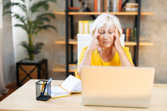 An elderly woman holding head with hands while sitting and working with a laptop indoors. Tired from work exhausted senior woman suffers from headache