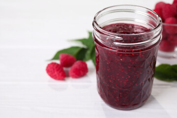 Delicious jam in glass jar and fresh raspberries on white wooden table. Space for text