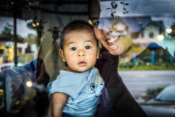 Happy young mother holding cute asian baby boy at coffee shop seen through window. Authentic lifestyle moments concept