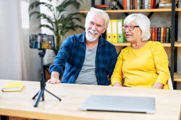 A cheerful senior couple of bloggers is recording a video, streaming online indoors. A gray-haired mature man and elderly woman sit at the desk in front of smartphone on tripod and laughth