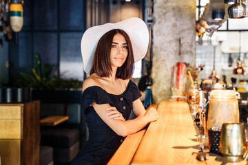 Fototapeta na wymiar beautiful woman in a black dress and a white wide-brimmed hat sitting at the bar in a cafe with a modern, trendy interior. Waiting for professional barista to prepare delicious, aromatic, fresh coffee