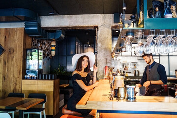 beautiful woman in black dress and a white wide-brimmed hat sitting at the bar in a cafe with a modern, trendy interior. Waiting for professional barista to prepare delicious, aromatic, fresh coffee