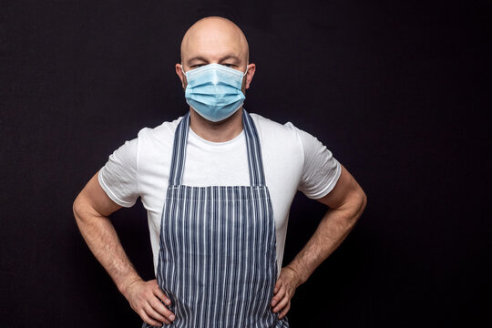 Professional Bald Butcher Or Fishmonger In White T-shirt And Black And White Apron On Dark Background. Man Wearing Blue Face Mask. Health Measures During COVID 19 Pandemic.