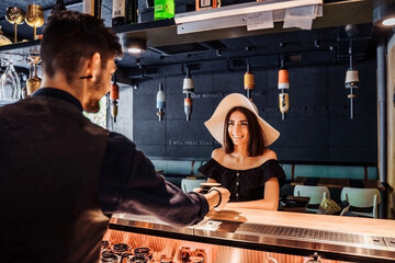 Professional bartender waiter with smile serving coffee to young woman. Regular visitor, sitting at the bar, communicating with interest with the barista. In favorite cafe with pleasant, yellow light