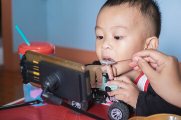 Cute little Asian baby boy accepting spoonful of food by his mom. Adorable son watching something on smartphone and playing with a toy car at home.