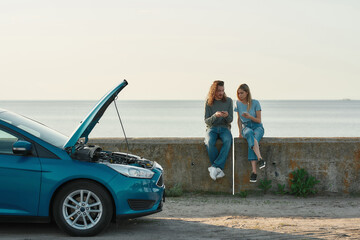 Outdoor shot of couple of travelers having problems with the car, man and woman sitting near broken...