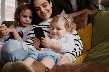 Mother and two daughters watching funny videos on smartphone sitting on sofa in modern lounge.