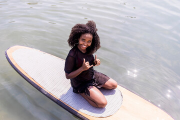 Young afro slim girl sitting on paddle Board in sea.