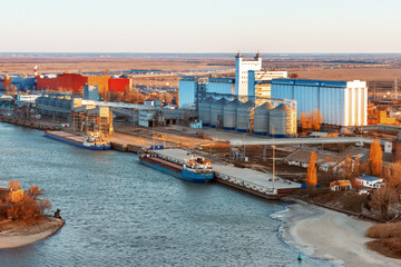 The large industrial building of a grain elevator placed on the bank of a river near a cargo berth...