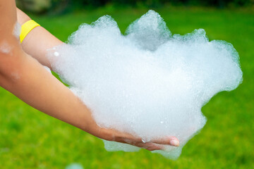 The girl's hands are holding a lot of soap suds, playing in the backyard on Summer day.Foam party at home yard with lot of shampoo for fun