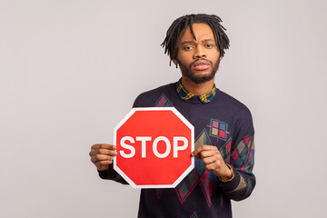 Concerned african guy with dreadlocks holding red stop sign in hands, attention, be careful concept, warning. Indoor studio shot isolated on gray background