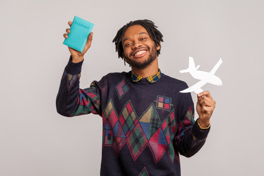 Cheerful Smiling African Guy With Dreadlocks Holding Passport And Paper Plane In Hands, Dreaming About Vacation Abroad, Adventures. Indoor Studio Shot Isolated On Gray Background