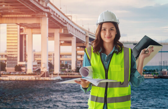 Female Civil Engineer With Computer And Blueprint Is Standing Infront City Tranportation Bridge Over River For City Development.