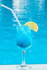 Blue lagoon in a glass with a straw and a slice of lemon. Summer sweet cocktail by the pool. Mixed alcohol drink. Beach party, vacation concept. Water surface.