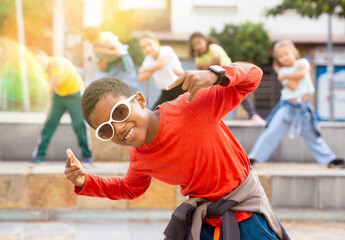 Portrait of confident african boy in sunglasses dancing hip-hop with group of tweens on summer city...