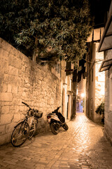 A narrow deserted street with bike and motorcycle in the medieval historical part of Trogir, Croatia lit by lanterns at night