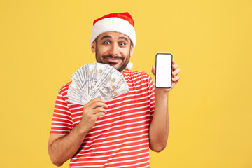 Happy bearded man in santa claus hat and red t-shirt holding fan of dollars and smartphone with empty blank, new year bonuses, advertising. Indoor studio shot isolated on yellow background