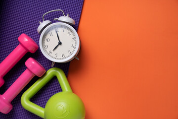 pink dumbbell, alarm clock, kettle bell and purple yoga mat on orange table background, fitness...