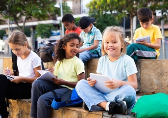 Positive preteen classmates preparing schoolwork, sitting on steps outside school in sunny spring...