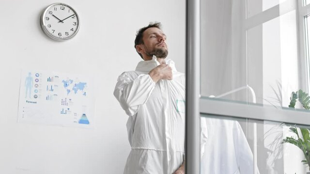 Panning of middle-aged Caucasian male virologist is standing in the office, zipping up white protective coverall and going away