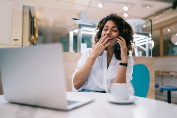 Cheerful ethnic woman laughing while talking on smartphone with friend in cafe