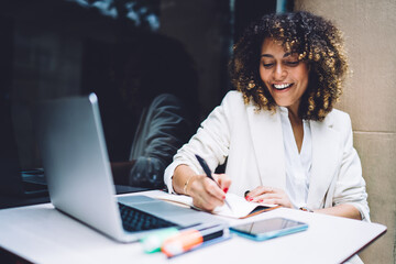 Satisfied ethnic woman working with laptop and daily planner at table