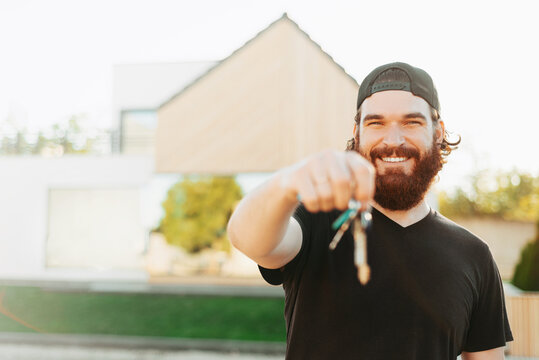 Cheerful Young Bearded Man Is Holding Some Keys And Pointing At Them Near His House