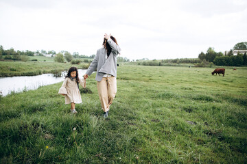 Stylish mother and daughter have fun outdoors in a field with green grass by the river when it rains