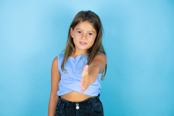 Young beautiful child girl over isolated blue background smiling friendly offering handshake as greeting and welcoming