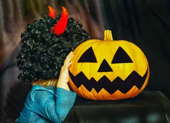 Foto op Plexiglas A child in a black wig with red horns hugs a pumpkin at a Halloween party. Adorable little girl hugs a big pumpkin on a pumpkin patch. Beautiful girl holding Jack o'lantern on a black background. © Наталья Микулич