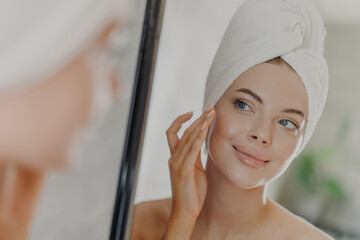 Beautiful woman touches face gently, looks at her reflection in mirror, enjoys softness of skin,...