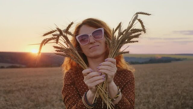 Red-haired woman in hat holding bouquet of spikelets of wheat in her hand in light of bright rays sun sunset lens flares slow motion at orange sunset sunny day. Woman walks in beautiful field blue sky
