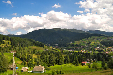 beautiful mountain landscape with bright clouds and sunlight in the sky, fluffy green spruces and indescribable beauty of the glade with residential buildings on the mountain slopes.