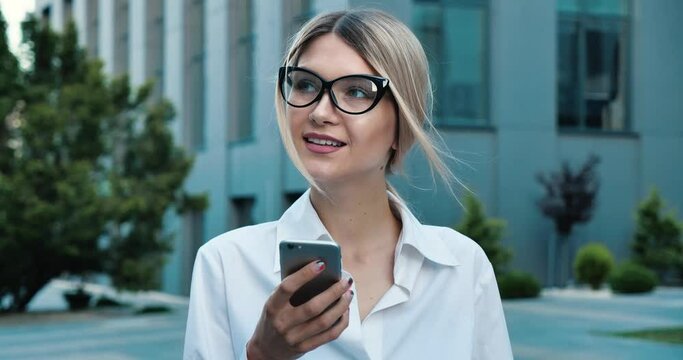 Young confident Businesswoman browsing Cellphone near modern Business Building. Light Wind blowing blonde Hair, having Charmingly Smile. Stylish Office Worker wearing clasiccal Dresscode, Glasses.