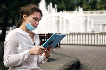 Obraz premium Young woman student in medical facial mask sits in quiet, deserted place in the park against the background of fountains. She looks at the tablet intently
