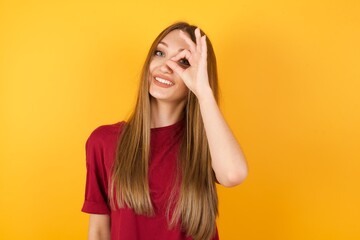 Beautiful Young beautiful caucasian girl wearing red t-shirt over isolated yellow background doing ok gesture with hand smiling, eye looking through fingers with happy face.