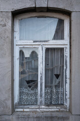 Fototapeta na wymiar Paintless and rotten window with torn net curtains. The street is reflected in the glass